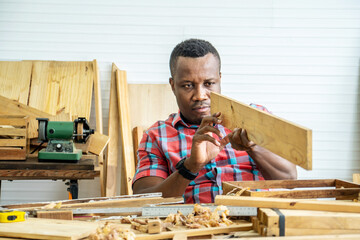 Wall Mural - Young carpenter african american man looking and choosing wood and using sandpaper to rub wooden plank at workshop table in carpenter wood factory