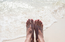 Women's Feet And A Sea Wave On A White Beach. Holiday Photo. Woman's Feet Near Wave On White Sand Beach. Sunny Afternoon At The Tropical Island.
