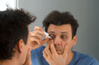 © Evgeniia - Man is dripping drops in his eyes in front of the mirror at home. He is looking at his reflection in the mirror. Eye fatigue, manager syndrome, exhaustion, stress, overwork, sleep disorder concepts.