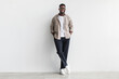 Handsome young black man in casual wear looking at camera and smiling, standing against white studio wall, copy space