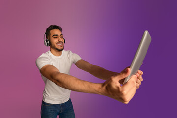 Poster - Handsome millennial Arab man chatting online, using tablet pc, wearing wireless headphones in neon light