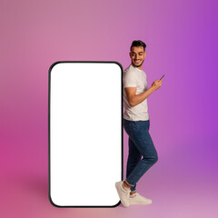 Poster - Happy young Arab man using mobile device while leaning on big smartphone with empty screen in neon light. Mockup