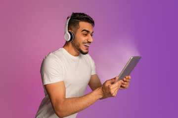 Wall Mural - Excited young Arab guy wearing headphones, looking at glowing tablet screen, overjoyed about huge sale in online store