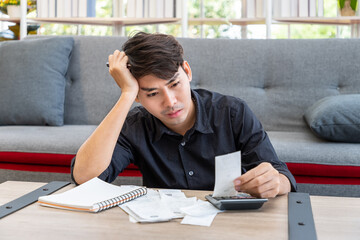 Serious Asian man calculating budget expenses receipt using calculator sitting in living room at home, personal finance concept
