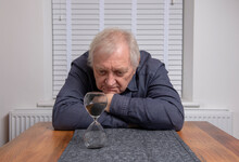 Senior Man Looking Into A Sand Hourglass Timer, Waiting.