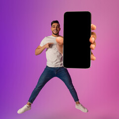 Poster - Handsome Arab guy showing big cellphone with empty black screen, jumping in air, offering space for mockup in neon light