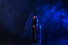 Electronic Cigarette, Vaping Device Mod, Stands In Vape Smoke And Vapor Illuminated By Blue Light.