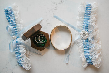 Wall Mural - Close up of wedding rings in a rustic wooden rings box with moss decoration inside Two blue bridal garters Bridal preparations flat lay White tablecloth background