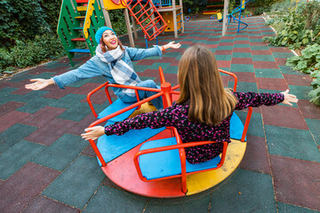 Two childish happy woman friends or teenage girl having fun on seesaw and carousel on a playground. Concept of psychology of the new generation and relations