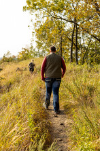 Young Couple Hiking Along Narrow Path In The Meadow In Autumn. Brilliant Autumn Colors On The Brush And Trees. Narrow Dirt Trail Through The Grass. 