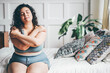 Curly haired overweight young woman in grey top and shorts with satisfaction on face accepts curvy body shape standing in stylish bedroom closeup