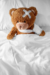 Wall Mural - medicine, healthcare and childhood concept - ill teddy bear toy with bandaged paw and medical patch on head lying in bed