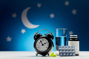 sleep disorder, bedtime and medicine concept - close up of alarm clock, glass of water, earplugs and sleeping pills over moon and night stars on blue background