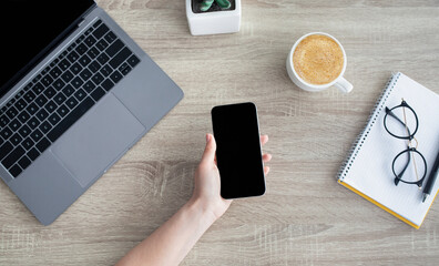 Poster - Female hand holding cellphone with blank black screen for your advertisement, sitting at workplace with laptop, top view