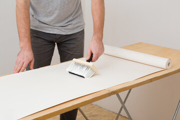 Young adult man hand using brush and applying glue on wallpaper sheet on wooden table. Closeup. Repair work of home.