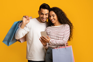Poster - Discount App. Cheerful Arab Couple Holding Smartphone And Colorful Paper Shopping Bags