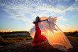© keleny - Beautiful young woman or girl in red dress and white wings on the sand on sunny day with blue sky. Angel model or dancer posing in photo shoot on dunes