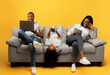 © Prostock-studio - Gadgets addiction. Young black family of three using different electronic devices, sitting on sofa on yellow background