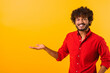 © Vadim Pastuh - Curly indian man wearing red casual style T-shirt, pointing at copy space on his palm, showing empty place for idea presentation, product advertising. Indoor studio shot isolated on orange background