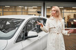 Successful young woman standing outside near white car on the background of car dealership and throwing up the car keys, looking at keys. Blonde playing, tossing her car keys into the air.