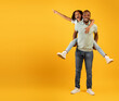 Joyful african american dad giving piggyback ride for daughter, girl pointing at free space on yellow background