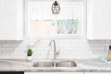 Kitchen Sink Detail Shot With A Marble Countertop, White Cabinets, Stainless Steel Faucet And Sink, And A Light Hanging In Front Of The Window.	