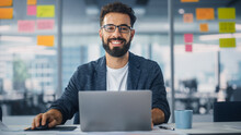 Modern Office: Portrait Of Stylish Hispanic Businessman Works On Laptop, Does Data Analysis And Creative Designer, Looks At Camera And Smiles. Digital Entrepreneur Works On E-Commerce Startup Project