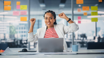 Poster - Young Successful Black Project Manager Dances and Smiles While Sitting at Her Desk. Beautiful Female Specialist Works on Laptop Computer in Creative Office, Celebrates Successful e-Commerce Project