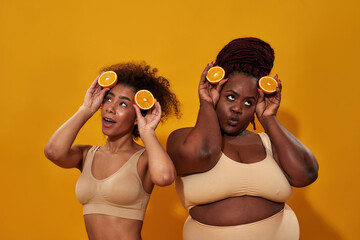 Two playful african american women looking silly, holding half of ripe orange, posing together isolated over orange background