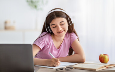 Wall Mural - Home schooling. Joyful Indian teenage girl in headphones taking notes during online class on laptop indoors