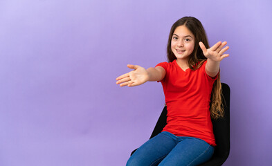 Wall Mural - Little caucasian girl sitting on a chair isolated on purple background presenting and inviting to come with hand