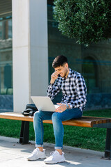 Handsome, smiling young guy talking on the phone while working with laptop and sitting on a bench on the street