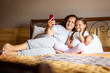 Smiling mother holding phone in hand with cute little daughter lying on double bed both looking at phone and talking with someone online wearing home clothes in daytime. Family time together.