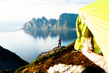 Hiker Admiring Mountains Standing On Top Of Cliff Beyond Camping Tent, Senja Island, Troms County, Norway, Scandinavia