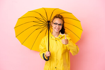 Wall Mural - Teenager Russian girl with rainproof coat and umbrella isolated on pink background giving a thumbs up gesture