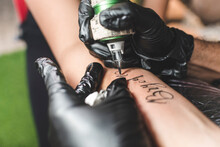 A Professional Tattoo Artist Uses A Rotary Tattoo Machine With A Round Liner Cartidge To Draw Cursive Names On A Female Client's Forearm.