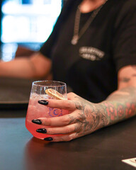 tattooed girl hand with black nails holding pink cocktail of vodka, tequila with ice and sliced oranges