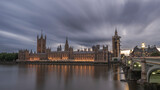 Fototapeta Londyn - The Houses of Parliament, London, England, illuminated as night falls and reflecting on the river Thames, with clouds streaking overhead