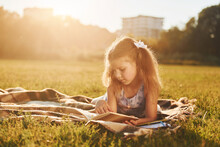 Little Girl Is Reading Book While Laying Down On The Grass Of Field Outdoors