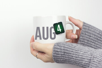 August 4th. Day 4 of month, Calendar date. Closeup of female hands in grey sweater holding cup of tea with month and calendar date on teabag label. Summer month, day of the year concept.