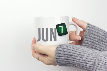 June 7th. Day 7 of month, Calendar date. Closeup of female hands in grey sweater holding cup of tea with month and calendar date on teabag label. Summer month, day of the year concept.