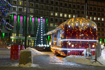 The red food truck decorated with garlands and lights stands on the square against the background of a Christmas tree