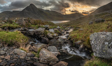 Epic Moody Autumn Sunset Landscape Image Of Llyn Ogwen And Tryfan In Snowdonia National Park With Stream And Rocks In Foreground