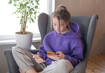 Girl in a lilac sweater sits with a phone in a chair near the window. Girl relaxing in chair using two smartphones.Communication, study at home, mobile apps, lockdown, technology, lifestyle, online