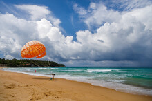 Opened Orange Parachute On The Beach In Thailand. The Man Offers To Go For A Parachute Ride
