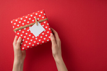 First person top view photo of saint valentine's day decorations female hands holding giftbox in red wrapping paper with heart pattern twine and pinned note on isolated red background with copyspace