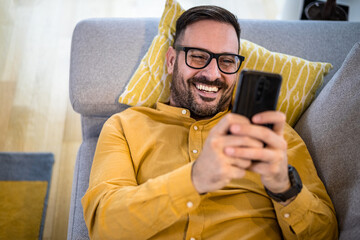 Cheerful young adult mature man happy smiling using smart phone resting lying down on sofa couch at home reading messages on mobile phone Guy with eyeglasses relaxing at home surfing net social media