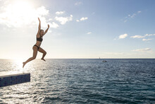 Young Woman Jumps From Diving Platform Into The Ocean Water Of The Caribbean Sea While On A Tropical Island Vacation On Cozumel, Mexico.