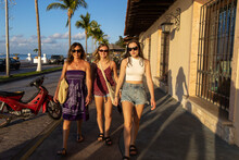 Three Women Walk Together On A Sidewalk In Mexico While On Vacation. Each Girl Is Wearing Sunglasses During The Golden Hour As The Sun Is Setting Over The Cozumel Beach Across The Street. 