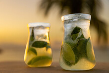 Fresh Muddled Mojitos With Rum, Lime And Mint In A Cold Glass With Ice. Perfect Refreshment During A Tropical Island Vacation With A Sunset And Palm Tree In The Background.